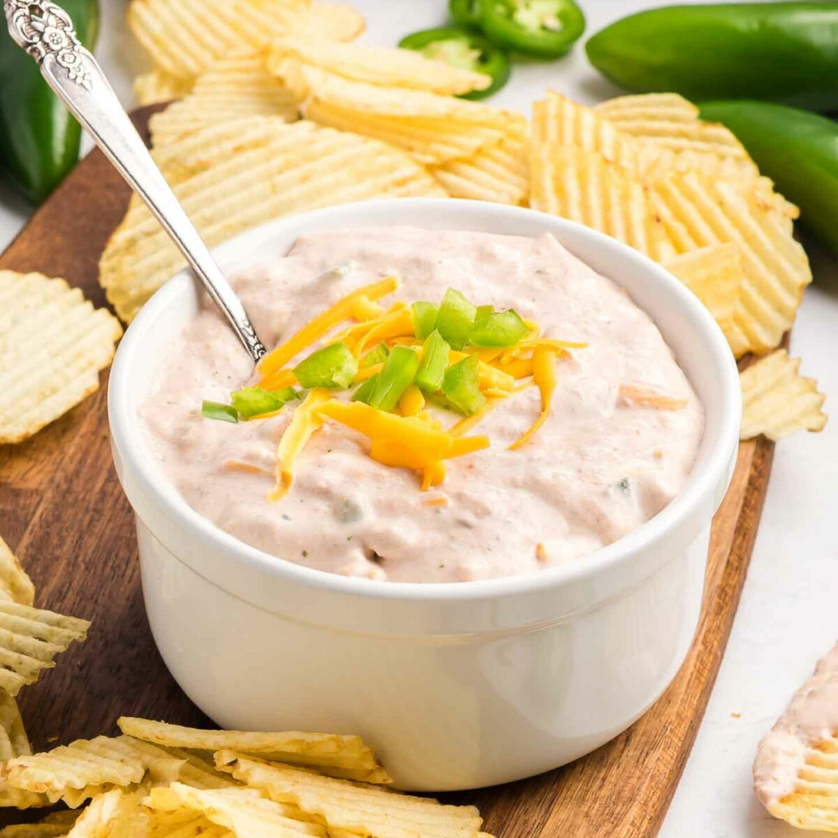 A white bowl of creamy dip topped with shredded cheese and diced green onions, surrounded by ridged potato chips on a wooden board with jalapeños and a striped cloth in the background.