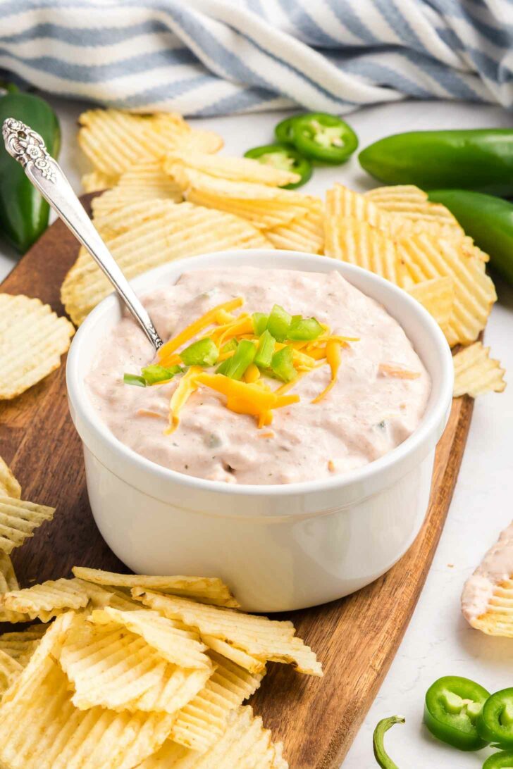 A white bowl of creamy dip topped with shredded cheese and diced green onions, surrounded by ridged potato chips on a wooden board with jalapeños and a striped cloth in the background.