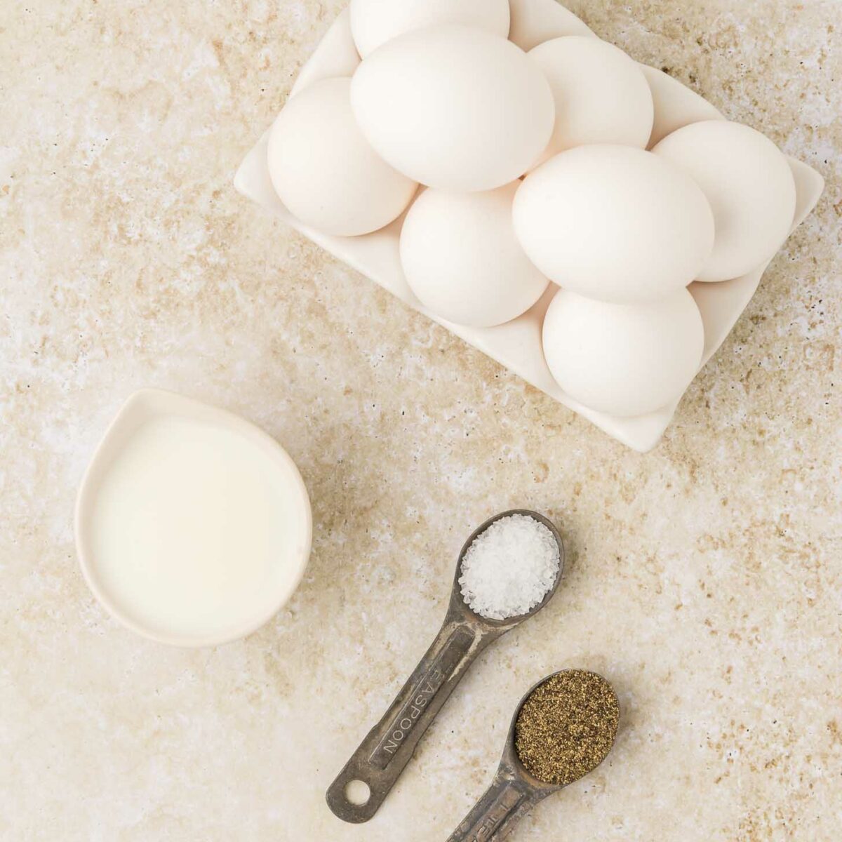 A carton of white eggs, a small pitcher of milk, and two measuring spoons filled with salt and ground black pepper on a light textured surface.
