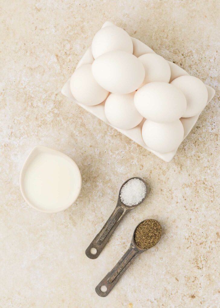 A carton of white eggs, a small pitcher of milk, and two measuring spoons filled with salt and ground black pepper on a light textured surface.
