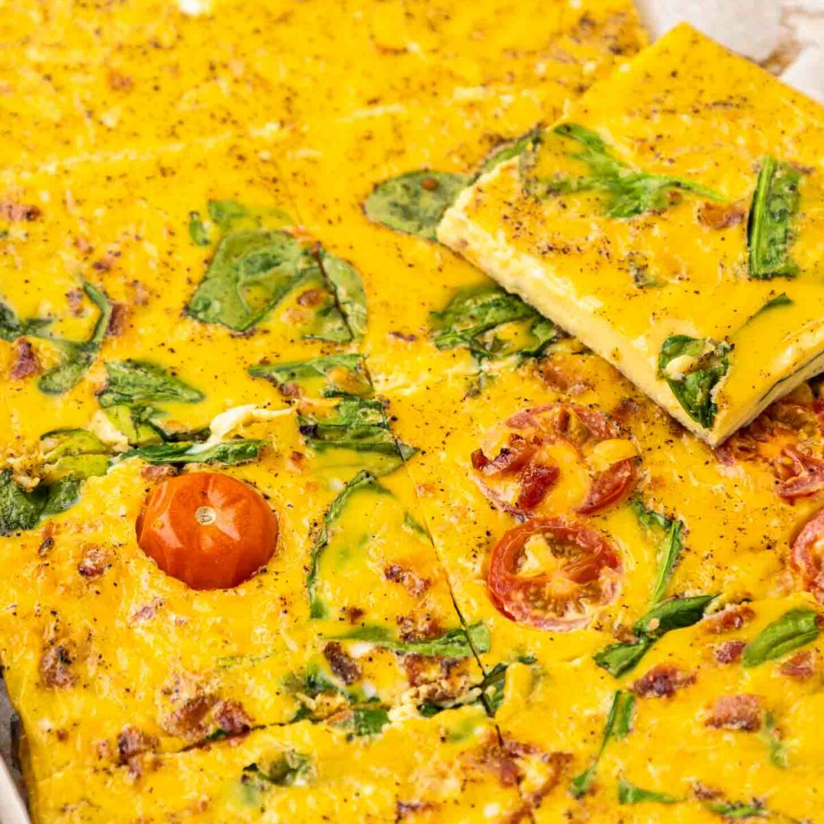 Sheet Pan Eggs cut into squares, topped with spinach leaves and sliced cherry tomatoes, with a golden-yellow, slightly speckled surface. One square is lifted to show the texture and ingredients inside.