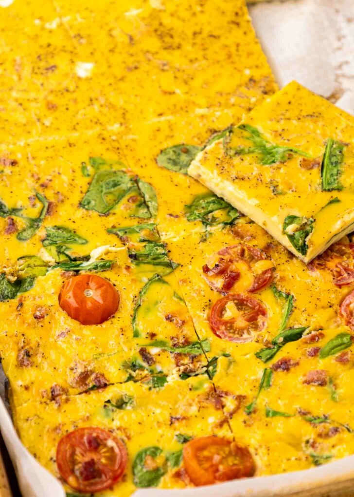 Sheet Pan Eggs cut into squares, topped with spinach leaves and sliced cherry tomatoes, with a golden-yellow, slightly speckled surface. One square is lifted to show the texture and ingredients inside.