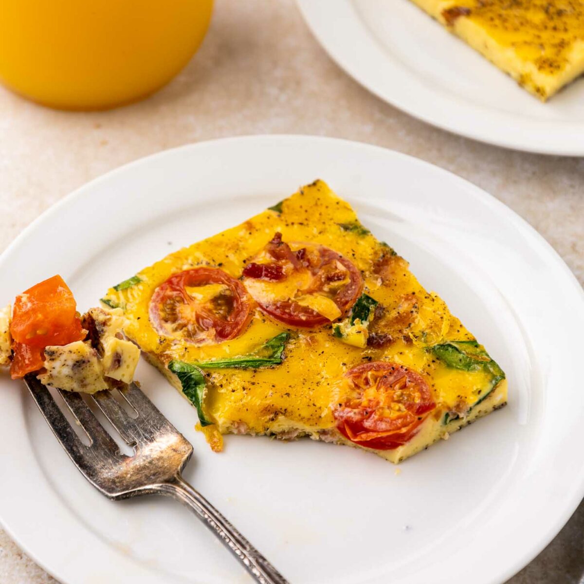 A slice of eggs with vegetables on top with cherry tomatoes and greens on a white plate, with a fork holding a bite. A glass of orange juice and another plate with more sheet pan eggs are in the background.