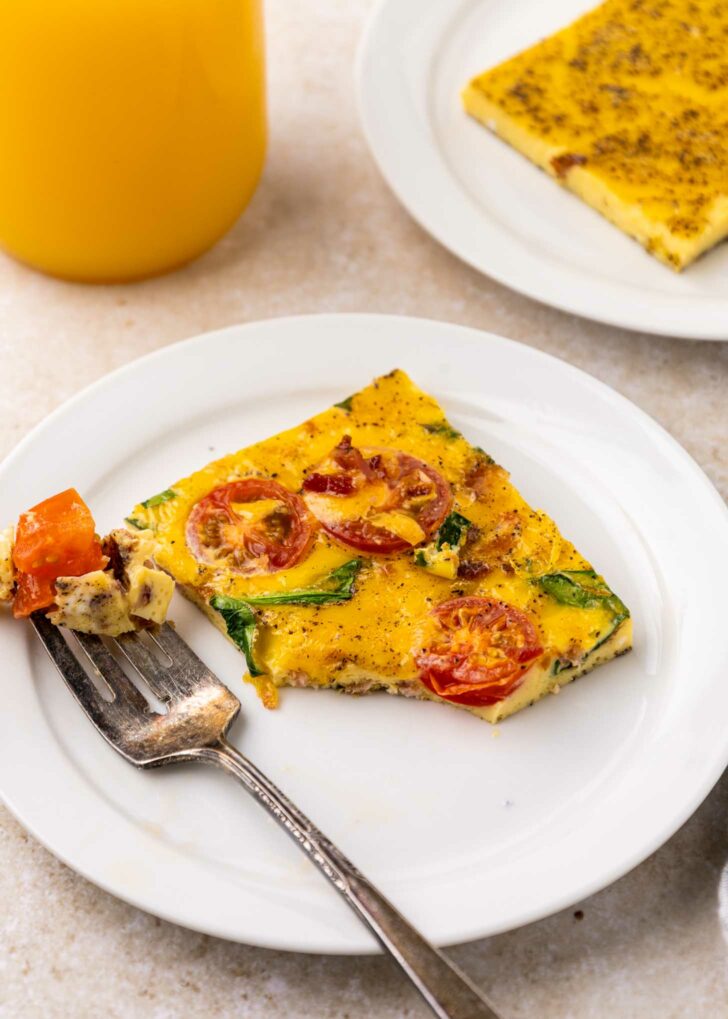 A slice of eggs with vegetables on top with cherry tomatoes and greens on a white plate, with a fork holding a bite. A glass of orange juice and another plate with more sheet pan eggs are in the background.