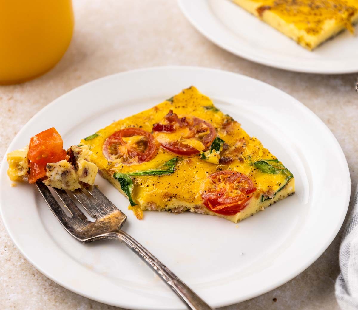 A slice of sheet pan eggs with tomatoes and greens on a white plate, next to a fork holding a bite. A glass of orange juice and a second plate are in the background.