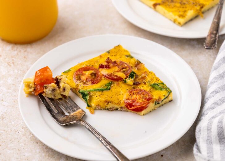 A slice of sheet pan eggs with tomatoes and greens on a white plate, next to a fork holding a bite. A glass of orange juice and a second plate are in the background.
