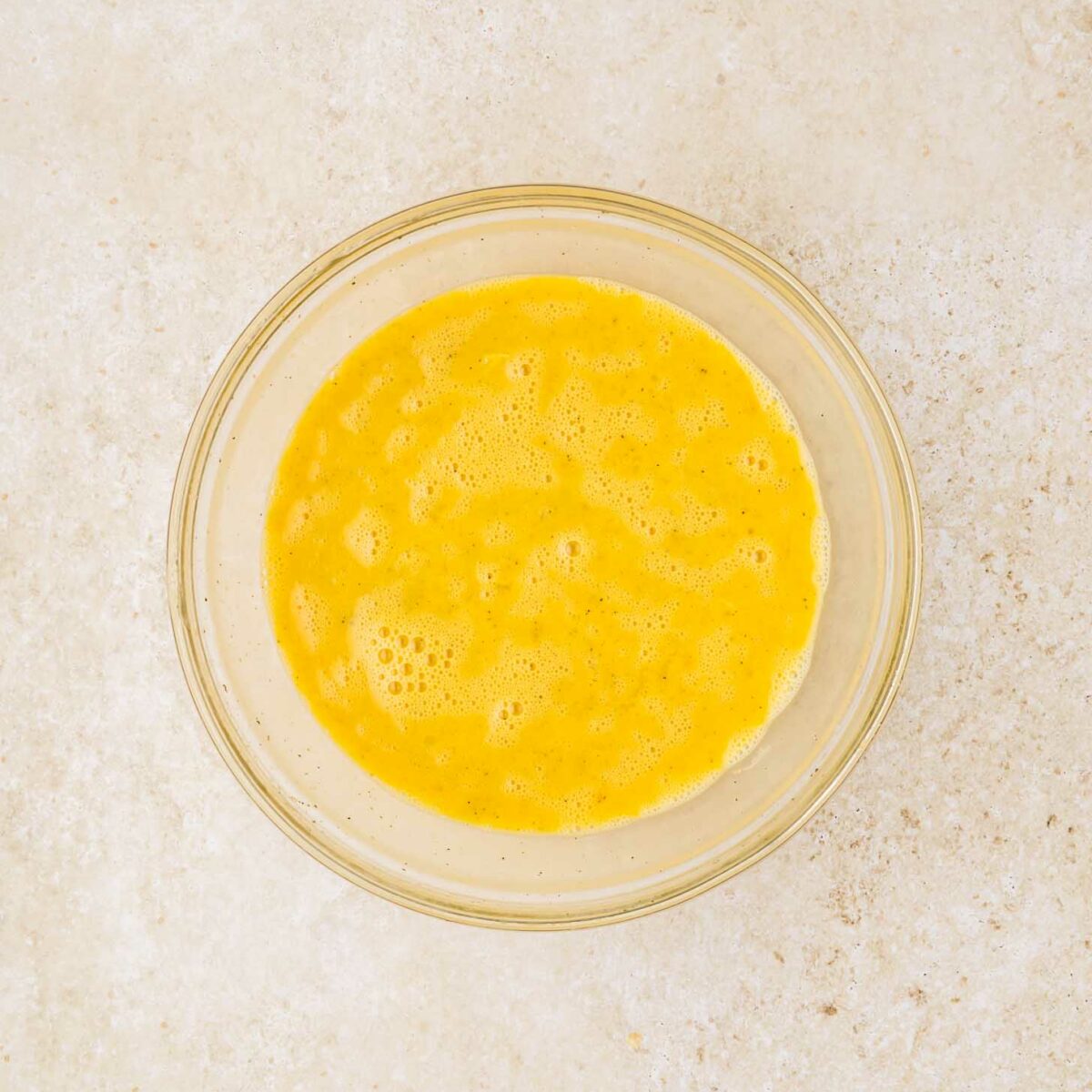 A glass bowl containing beaten eggs sits on a light-colored textured surface.