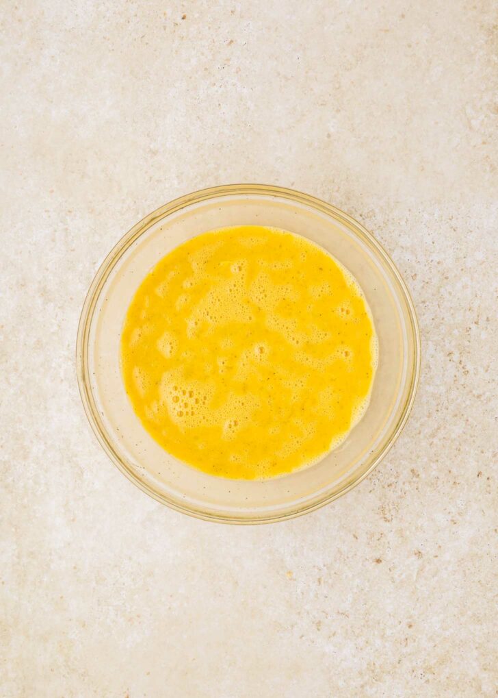 A glass bowl containing beaten eggs sits on a light-colored textured surface.
