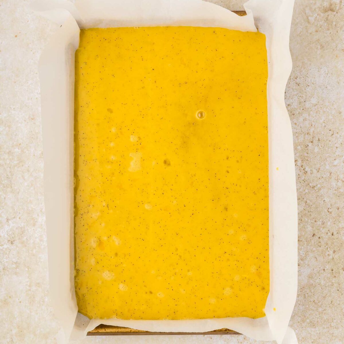 A rectangular baking pan lined with white parchment paper is filled with whisked eggs, speckled with tiny dots, and set on a light beige surface.