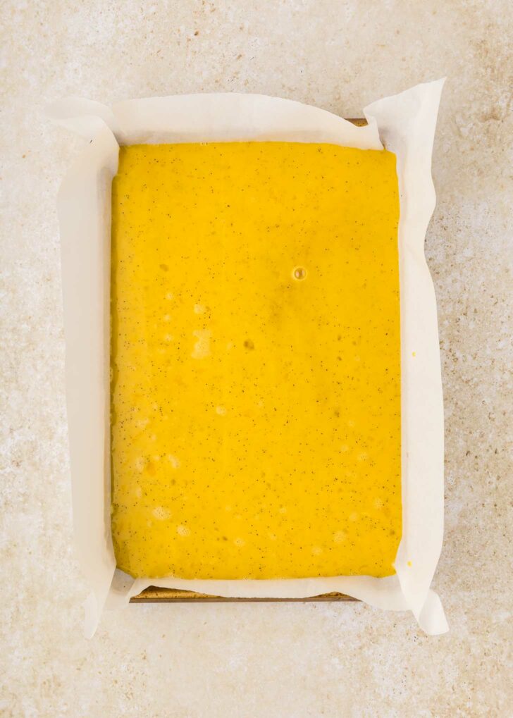 A rectangular baking pan lined with white parchment paper is filled with whisked eggs, speckled with tiny dots, and set on a light beige surface.