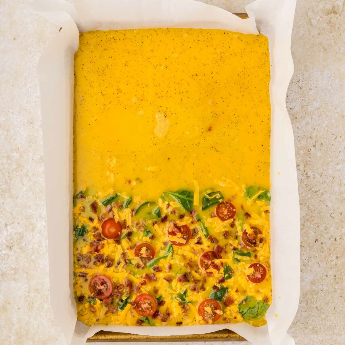 Rectangular baking pan lined with parchment paper, filled with an uncooked egg mixture. The bottom half has visible spinach, tomatoes, and cheese, while the top half is only yellow liquid.