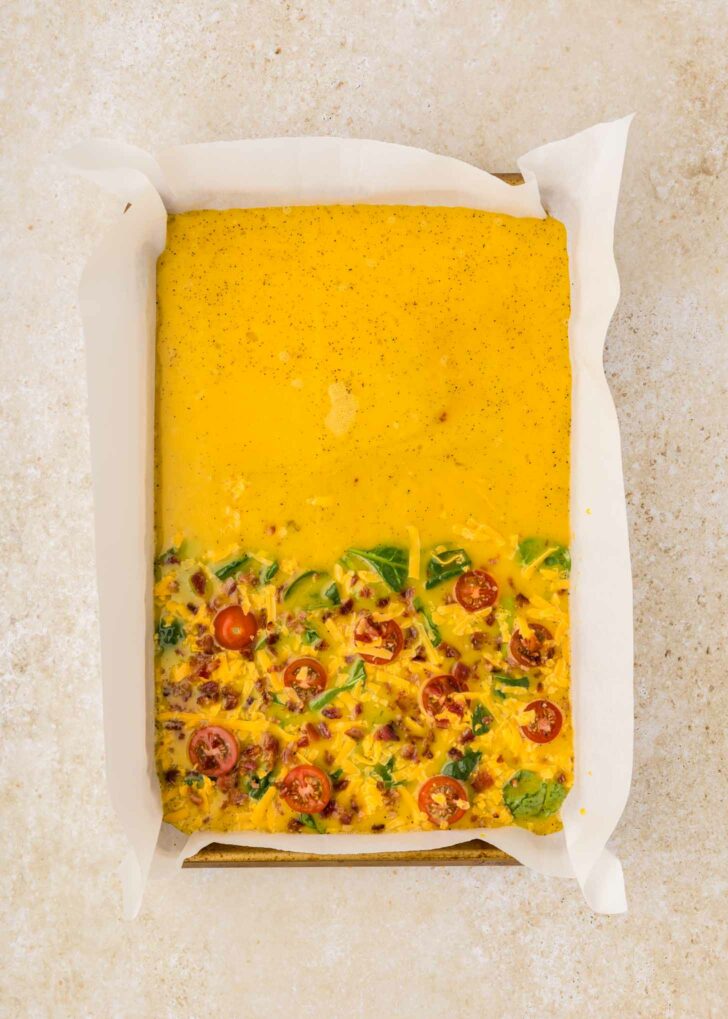 Rectangular baking pan lined with parchment paper, filled with an uncooked egg mixture. The bottom half has visible spinach, tomatoes, and cheese, while the top half is only yellow liquid.