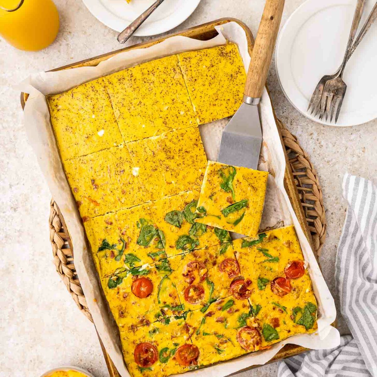 A baking tray with parchment paper holds a sliced, baked eggs with vegetables. A spatula lifts a piece. Plates, forks, orange juice, and a bowl of shredded cheese are nearby on a light countertop.