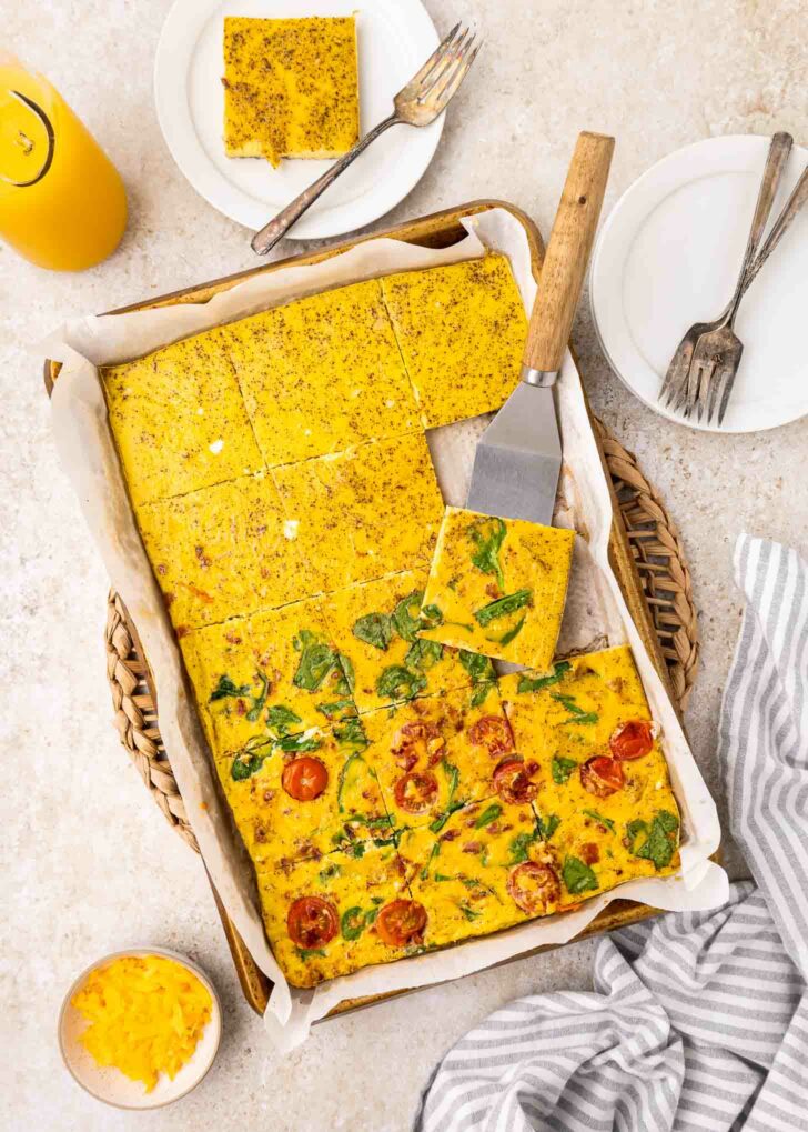 A baking tray with parchment paper holds a sliced, baked eggs with vegetables. A spatula lifts a piece. Plates, forks, orange juice, and a bowl of shredded cheese are nearby on a light countertop.