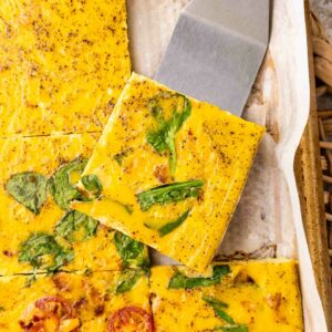 A close-up of baked egg squares with spinach and tomatoes, cut into pieces on parchment paper. A spatula lifts one square, showing the colorful vegetables and seasoning on the eggs surface.