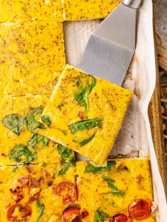 A close-up of baked egg squares with spinach and tomatoes, cut into pieces on parchment paper. A spatula lifts one square, showing the colorful vegetables and seasoning on the eggs surface.
