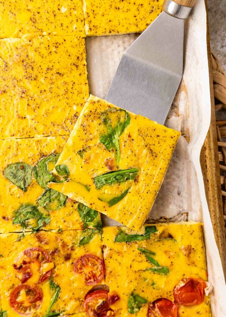 A close-up of baked egg squares with spinach and tomatoes, cut into pieces on parchment paper. A spatula lifts one square, showing the colorful vegetables and seasoning on the eggs surface.