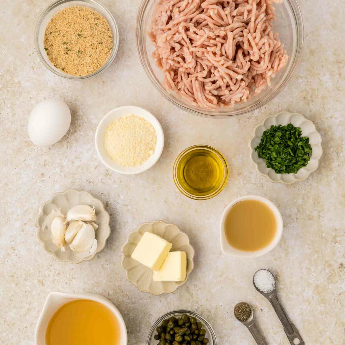Top-down view of ingredients in small bowls and dishes: ground chicken, breadcrumbs, egg, grated cheese, minced parsley, olive oil, butter, garlic cloves, chicken broth, capers, salt, and pepper.