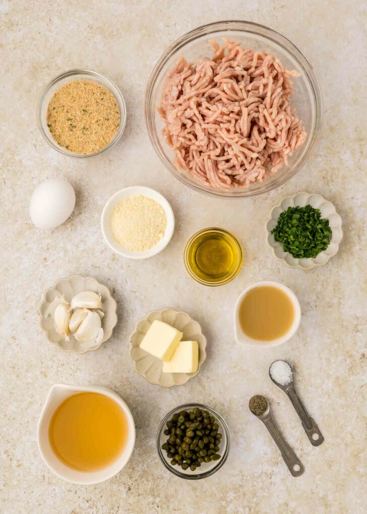 Top-down view of ingredients in small bowls and dishes: ground chicken, breadcrumbs, egg, grated cheese, minced parsley, olive oil, butter, garlic cloves, chicken broth, capers, salt, and pepper.