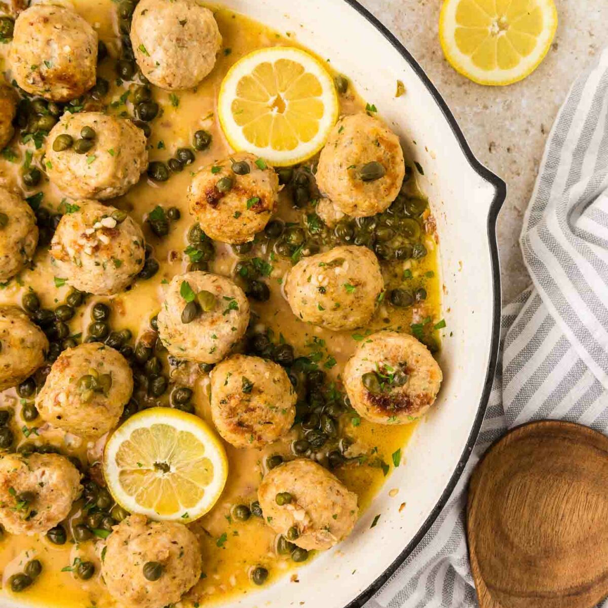 Chicken piccata meatballs in a creamy lemon-caper sauce are served in a white skillet, garnished with lemon slices and parsley. A wooden spoon and striped towel are nearby, with two lemon slices on the countertop.