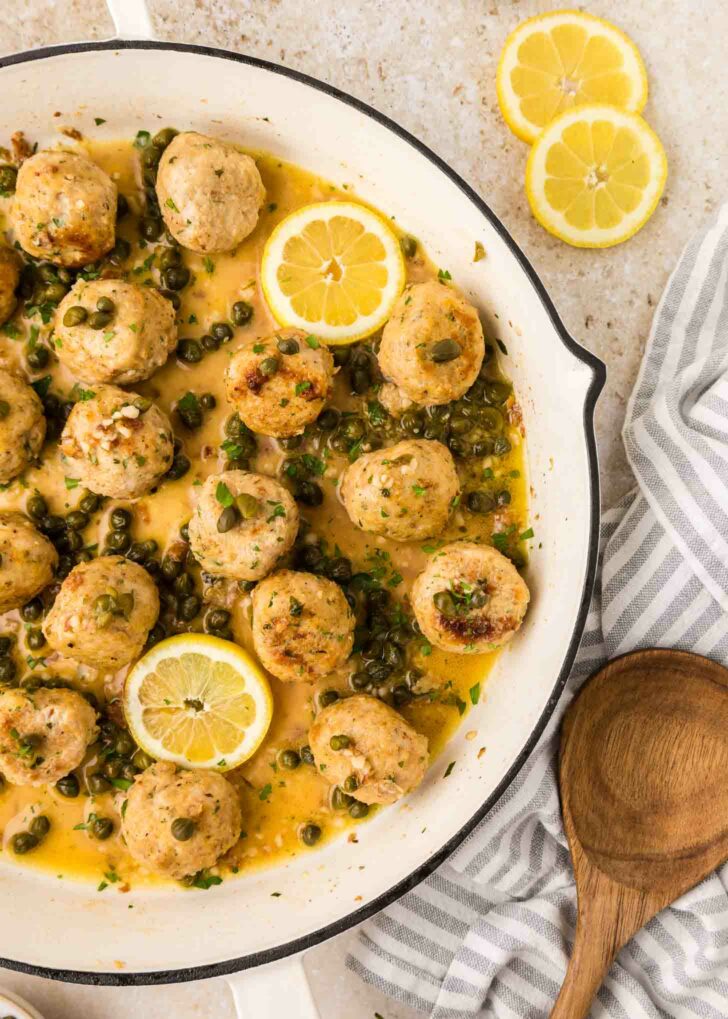 Chicken piccata meatballs in a creamy lemon-caper sauce are served in a white skillet, garnished with lemon slices and parsley. A wooden spoon and striped towel are nearby, with two lemon slices on the countertop.