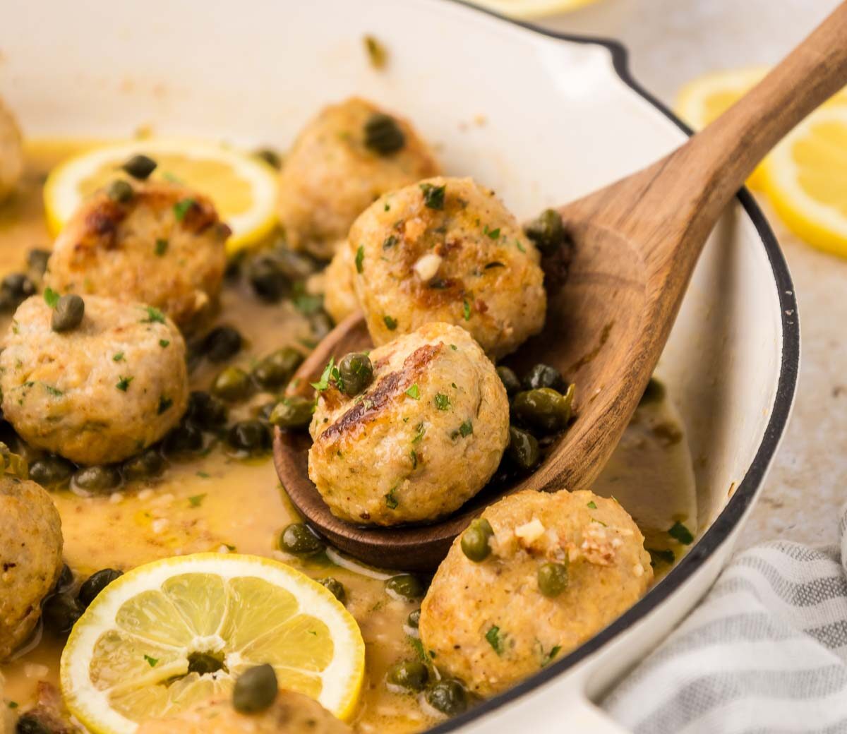 A wooden spoon lifts two chicken meatballs from a pan filled with more meatballs, capers, lemon slices, and a light sauce. A striped cloth is visible beside the pan.