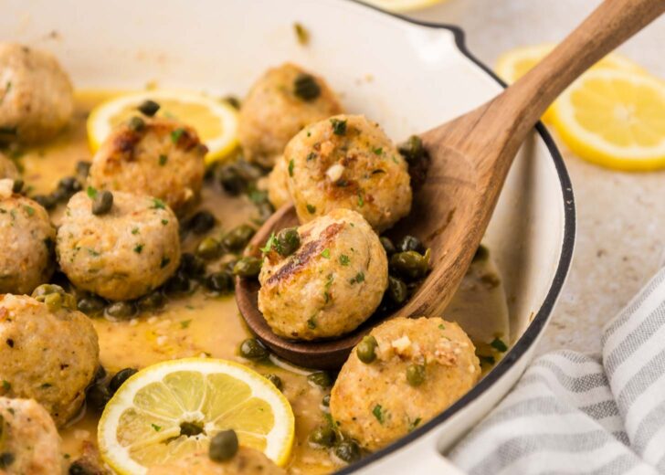 A wooden spoon lifts two chicken meatballs from a pan filled with more meatballs, capers, lemon slices, and a light sauce. A striped cloth is visible beside the pan.