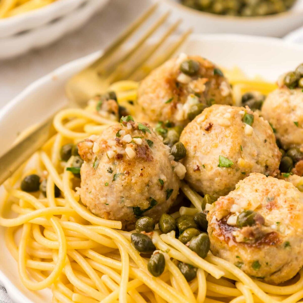 A plate of spaghetti topped with several browned meatballs, garnished with chopped herbs and capers, with a gold fork resting on the side. In the background, there is another bowl of pasta and a small dish of capers.