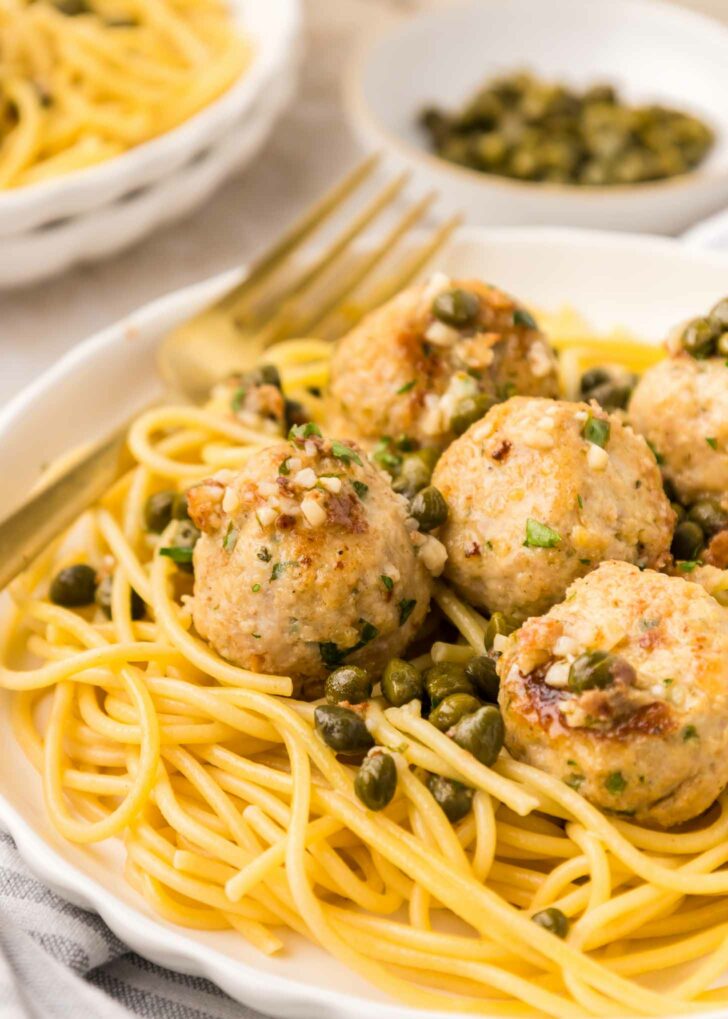 A plate of spaghetti topped with several browned meatballs, garnished with chopped herbs and capers, with a gold fork resting on the side. In the background, there is another bowl of pasta and a small dish of capers.