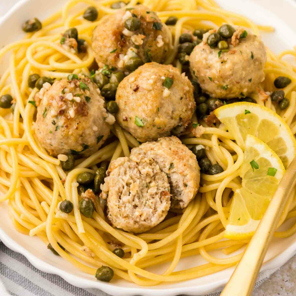 A plate of spaghetti topped with five chicken piccata meatballs, capers, and fresh herbs, garnished with lemon slices. One meatball is cut in half to show the inside. A gold fork rests on the side of the plate.