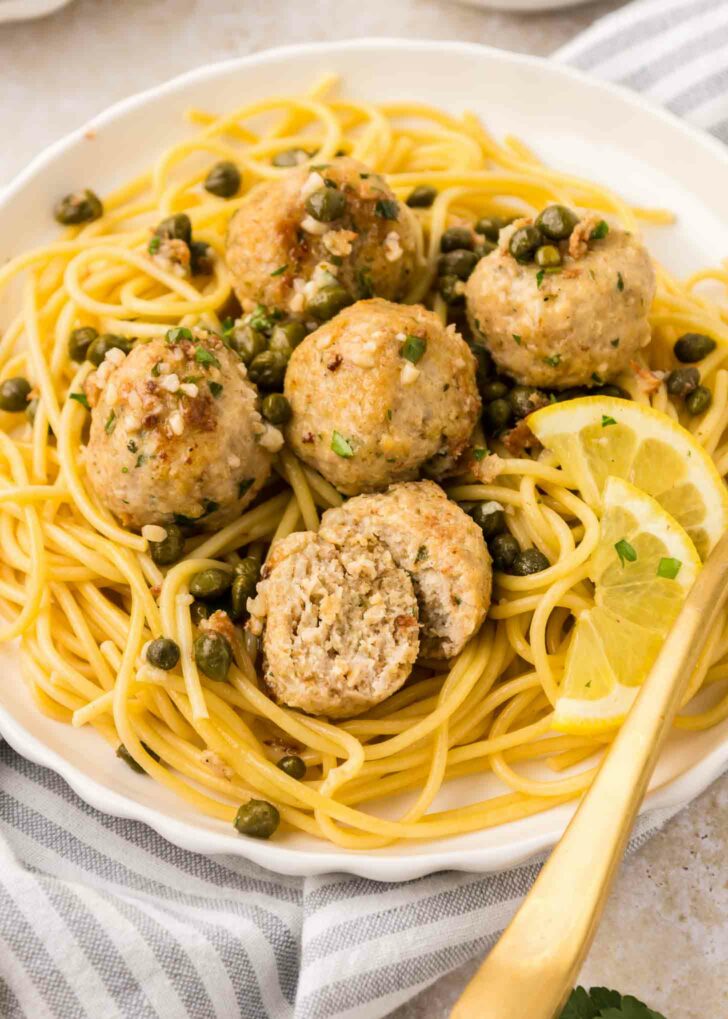 A plate of spaghetti topped with five chicken piccata meatballs, capers, and fresh herbs, garnished with lemon slices. One meatball is cut in half to show the inside. A gold fork rests on the side of the plate.