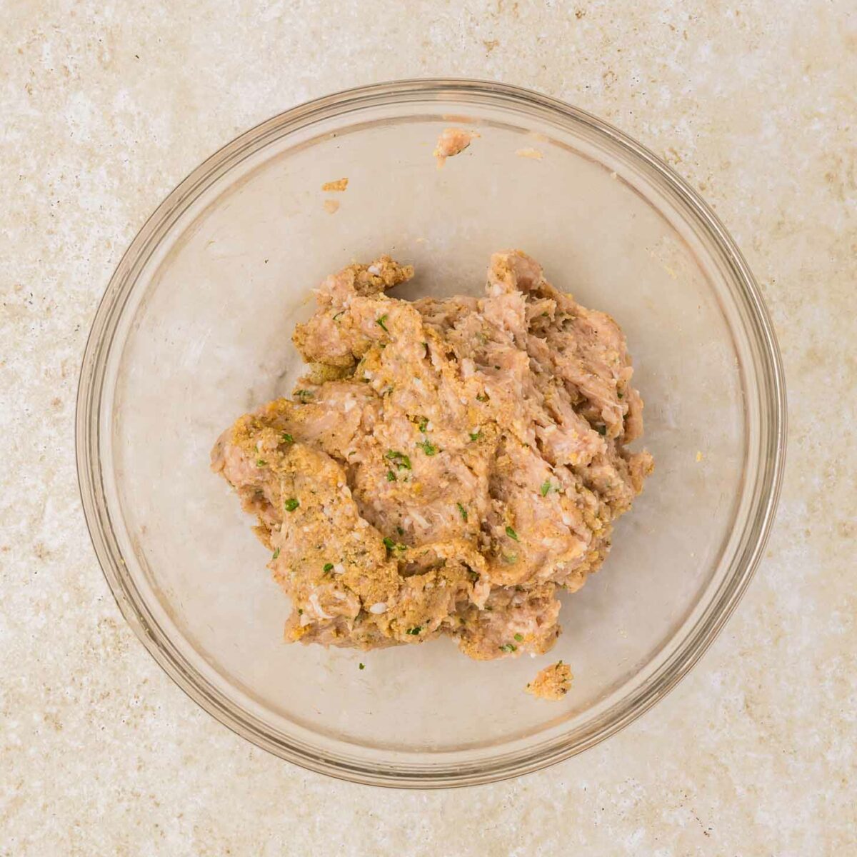 A clear glass bowl containing a mixture of ground chicken and spices sits on a light-colored countertop.