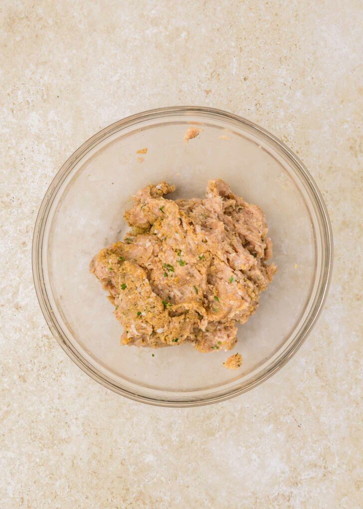 A clear glass bowl containing a mixture of ground chicken and spices sits on a light-colored countertop.