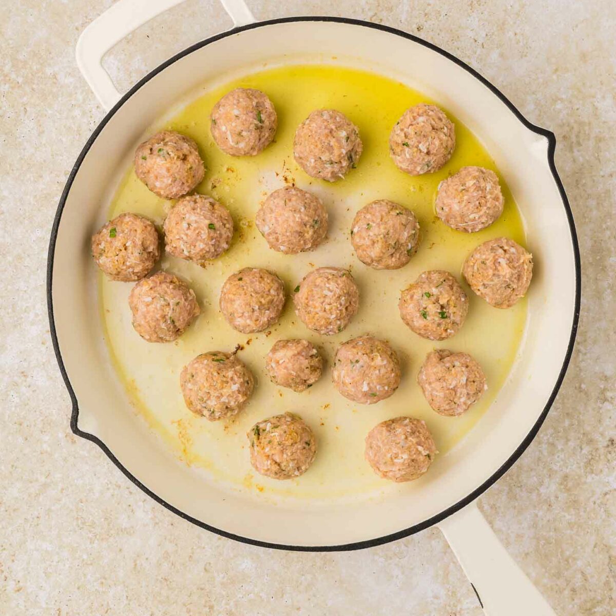 A white skillet filled with uncooked meatballs sits on a light-colored countertop. The meatballs are evenly spaced and surrounded by a small amount of oil.