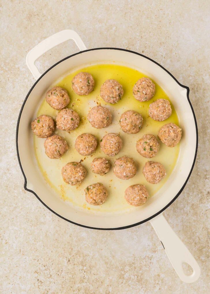 A white skillet filled with uncooked meatballs sits on a light-colored countertop. The meatballs are evenly spaced and surrounded by a small amount of oil.