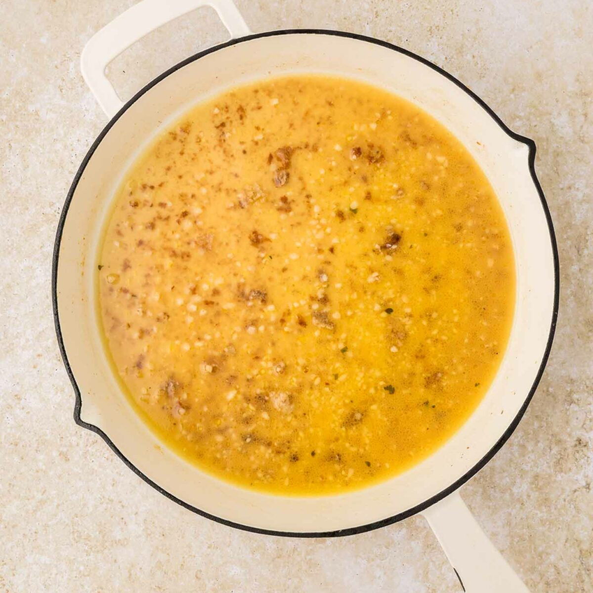 A white enameled pot filled with a yellow broth containing small bits of browned food, sitting on a light-colored, textured surface.