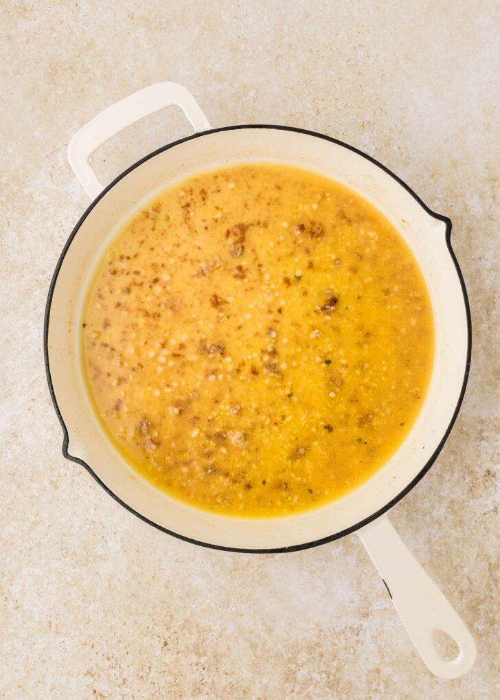 A white enameled pot filled with a yellow broth containing small bits of browned food, sitting on a light-colored, textured surface.