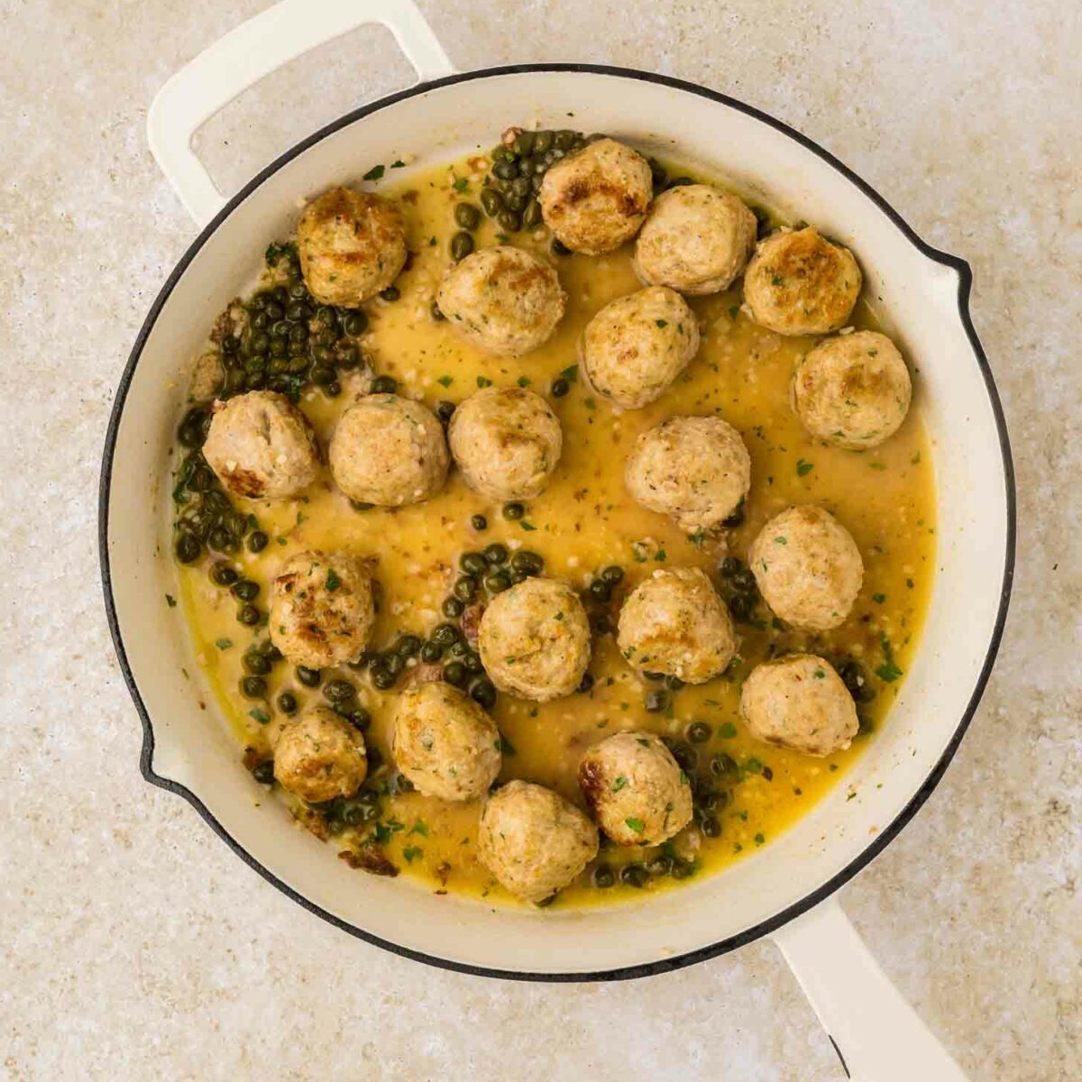 A white skillet filled with meatballs in a creamy yellow sauce, garnished with capers and herbs, placed on a light-colored surface.