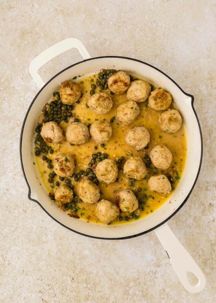 A white skillet filled with meatballs in a creamy yellow sauce, garnished with capers and herbs, placed on a light-colored surface.
