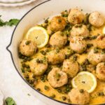 A white skillet filled with chicken piccata meatballs in a lemon-caper sauce, garnished with parsley and lemon slices. Plates and cutlery are visible in the background.