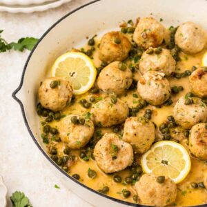 A white skillet filled with chicken piccata meatballs in a lemon-caper sauce, garnished with parsley and lemon slices. Plates and cutlery are visible in the background.