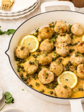 A white skillet filled with chicken piccata meatballs in a lemon-caper sauce, garnished with parsley and lemon slices. Plates and cutlery are visible in the background.