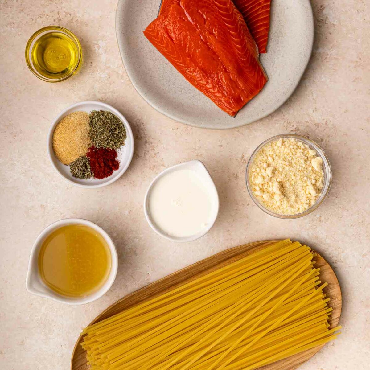 Uncooked spaghetti, raw salmon fillets on a plate, bowls of olive oil, spices, grated cheese, cream, broth, and garlic powder arranged on a light-colored countertop.