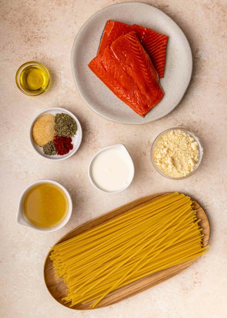 Uncooked spaghetti, raw salmon fillets on a plate, bowls of olive oil, spices, grated cheese, cream, broth, and garlic powder arranged on a light-colored countertop.