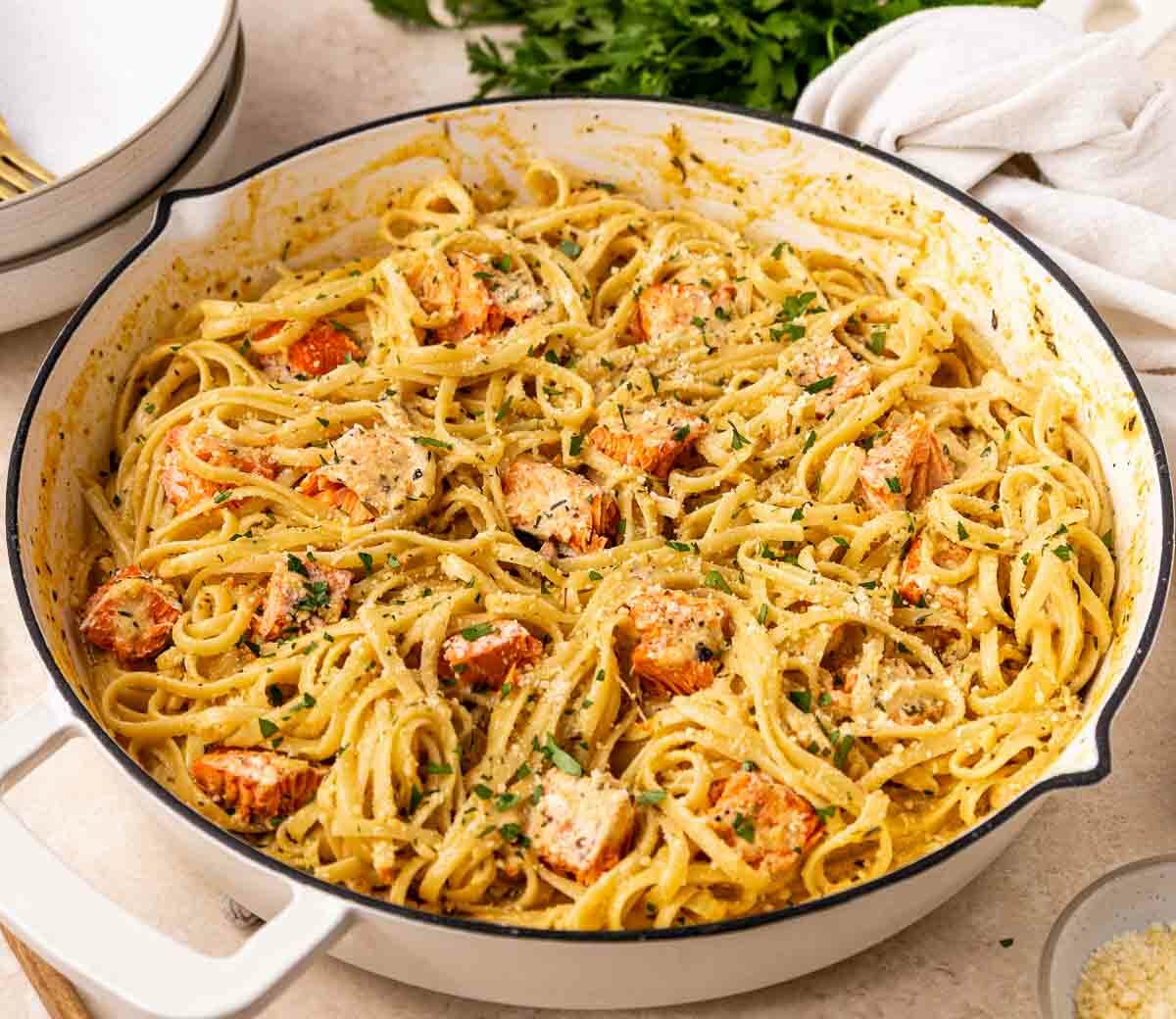 A white pan filled with creamy fettuccine pasta, pieces of cooked salmon, and chopped herbs, set on a countertop with a white cloth, plates, and utensils nearby.