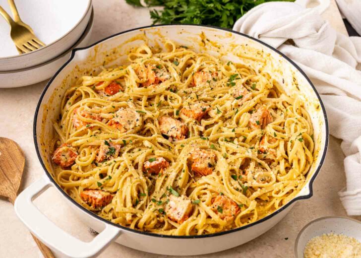 A white pan filled with creamy fettuccine pasta, pieces of cooked salmon, and chopped herbs, set on a countertop with a white cloth, plates, and utensils nearby.