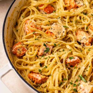 A close-up of creamy fettuccine pasta mixed with chunks of salmon and garnished with chopped parsley, served in a white skillet.
