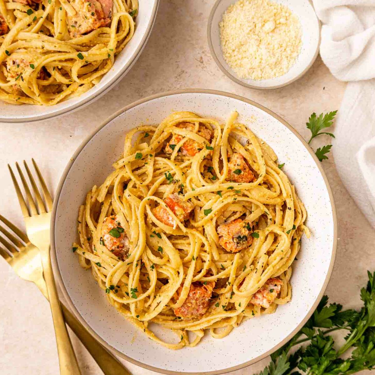 A bowl of creamy pasta with salmon chunks and herbs, garnished with parsley. Another bowl, a small dish of grated cheese, two gold forks, and green herbs are nearby on a light-colored surface.