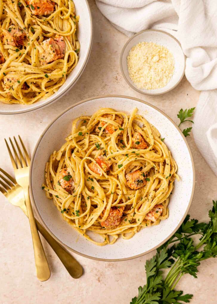 A bowl of creamy pasta with salmon chunks and herbs, garnished with parsley. Another bowl, a small dish of grated cheese, two gold forks, and green herbs are nearby on a light-colored surface.