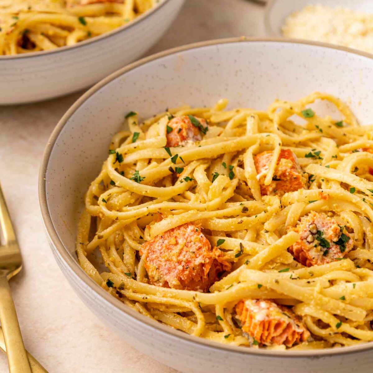 A close-up of a bowl of creamy pasta with chunks of salmon, garnished with chopped herbs. Another bowl and a plate of grated cheese are in the background, alongside gold-colored utensils.